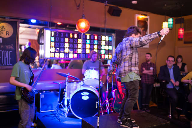 Youth performing on stage with a microphone at a live music event, accompanied by a band featuring a drummer and guitarist, colorful lighting in a lively venue, highlighting community engagement in music education.